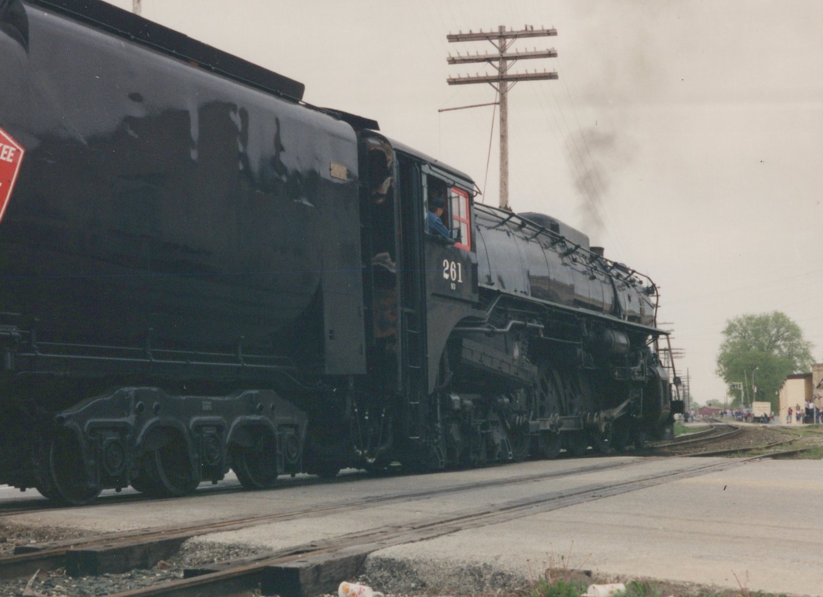 A black steam locomotive numbered 261 is moving on railroad tracks at a grade crossing with spectators watching.