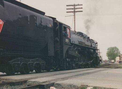 A black steam locomotive numbered 261 is moving on railroad tracks at a grade crossing with spectators watching.