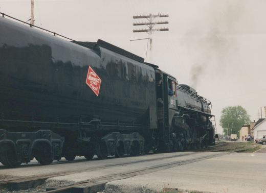 A black steam locomotive with the number 261 and a red diamond herald for the Chicago, Milwaukee, St. Paul and Pacific railroad is moving on tracks alongside a street, with smoke rising from its stack and people watching from the side.