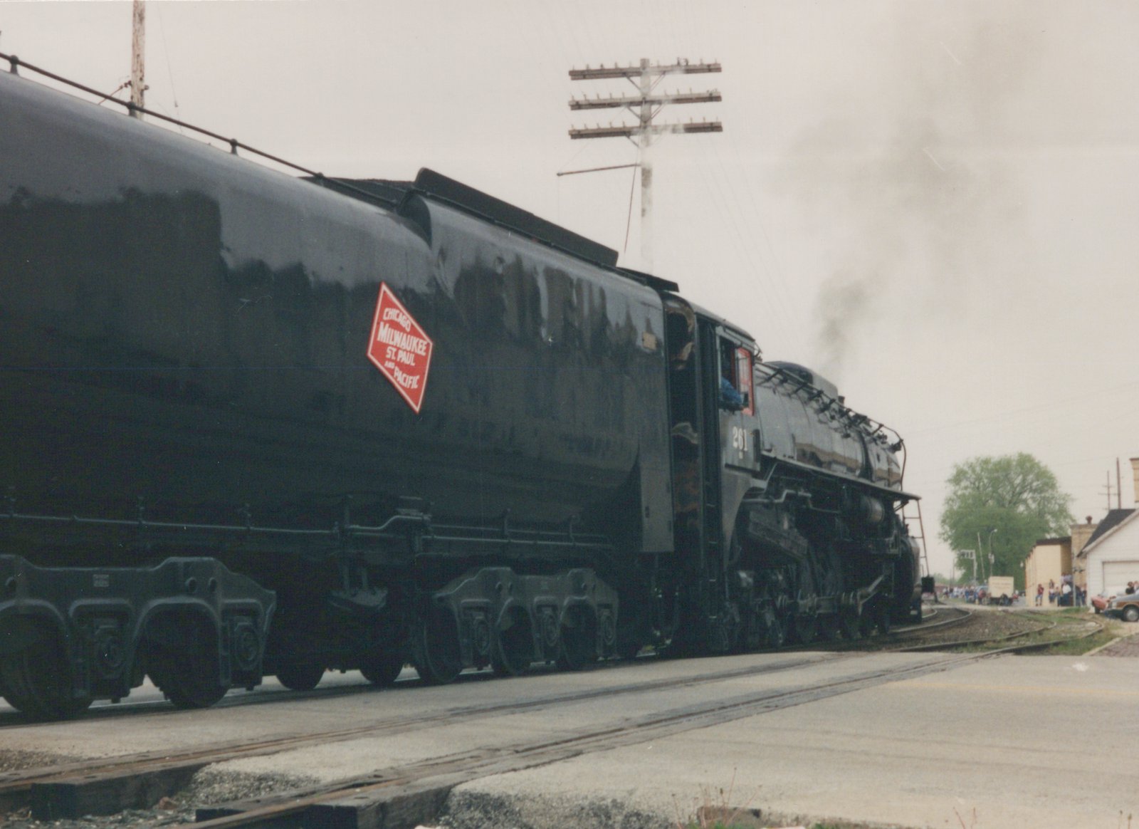 A black steam locomotive with the number 261 and a red diamond herald for the Chicago, Milwaukee, St. Paul and Pacific railroad is moving on tracks alongside a street, with smoke rising from its stack and people watching from the side.