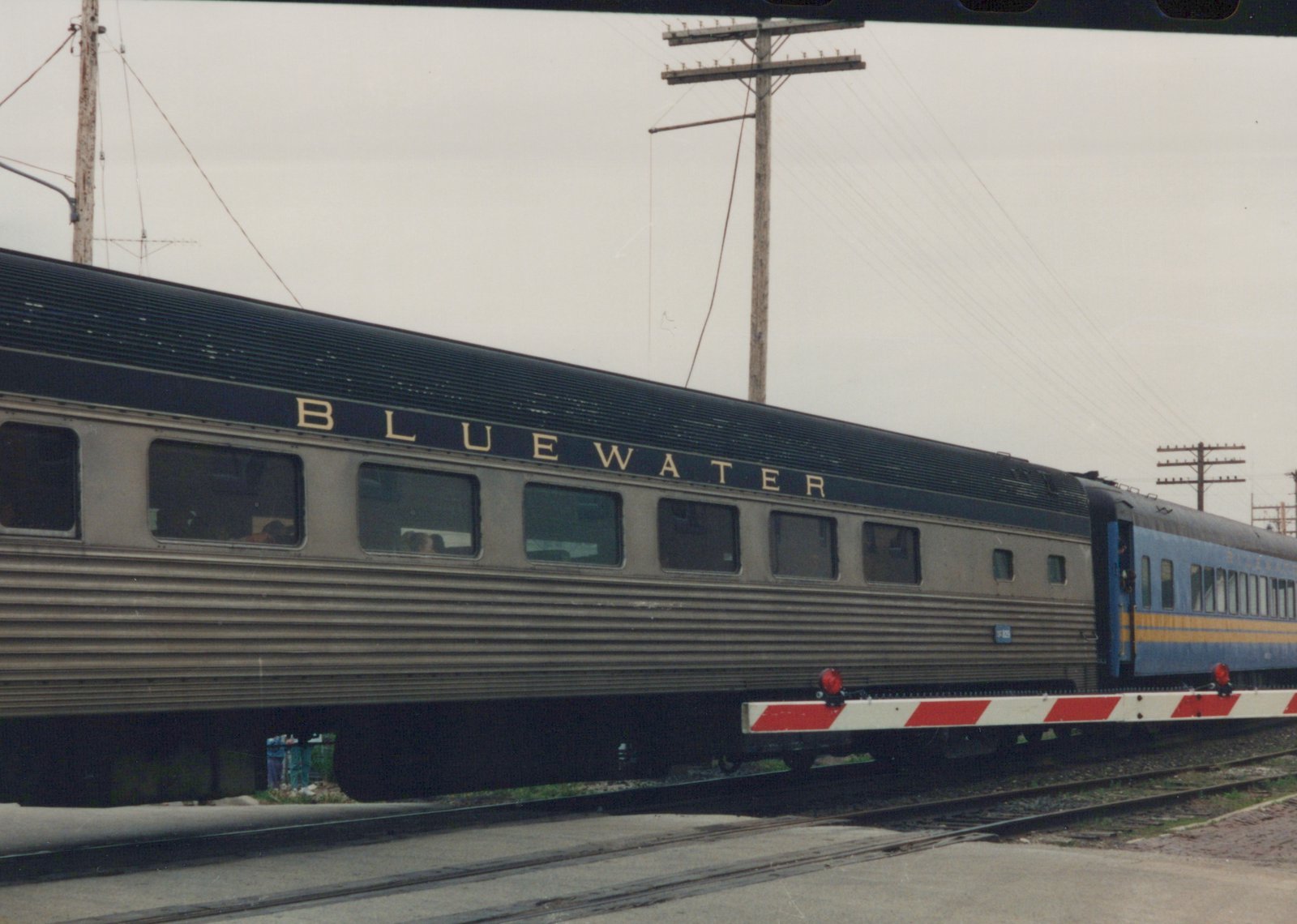 Side view of an Amtrak Blue Water passenger train with a gray heavyweight car numbered 325, stopped at a grade crossing with a red and white striped crossing gate.