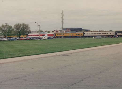 A wide shot of a grassy field with a line of vintage railroad cars in the background, several people walking around, and a row of parked cars from the 1970s or 1980s in the mid-ground.