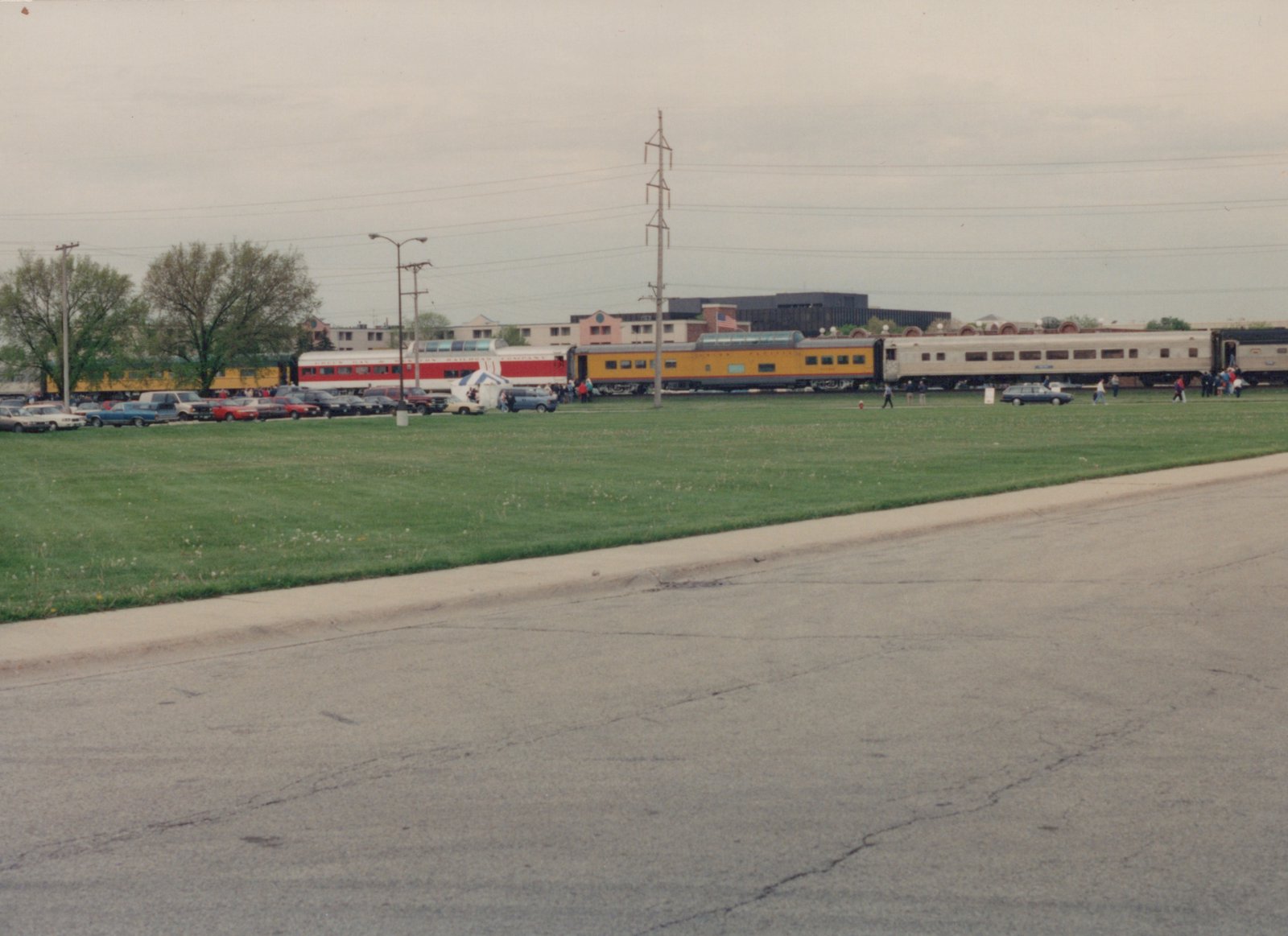 A wide shot of a grassy field with a line of vintage railroad cars in the background, several people walking around, and a row of parked cars from the 1970s or 1980s in the mid-ground.