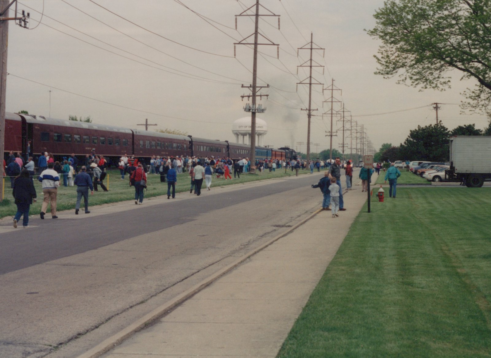 A large crowd gathers on a sidewalk to watch a vintage Milwaukee Road steam train travel past a water tower marked 'City of Des Plaines' under an overcast sky.