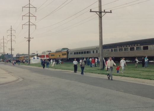 A long train with 'BLUEWATER' written on its side is parked beside a road where many people are walking and standing, with a large tent and power lines visible in the background.