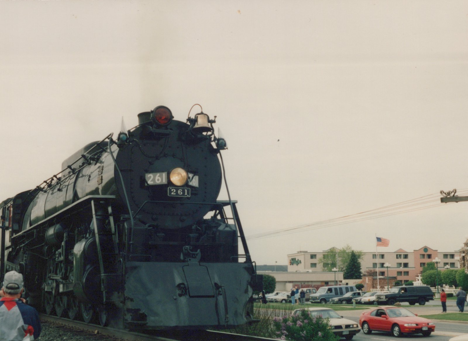 A black Canadian National steam locomotive numbered 261 is stopped on a street-level railroad crossing, with 1990s cars and onlookers in the background under an overcast sky.