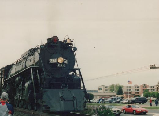 A black Canadian National steam locomotive numbered 261 is stopped on a street-level railroad crossing, with 1990s cars and onlookers in the background under an overcast sky.