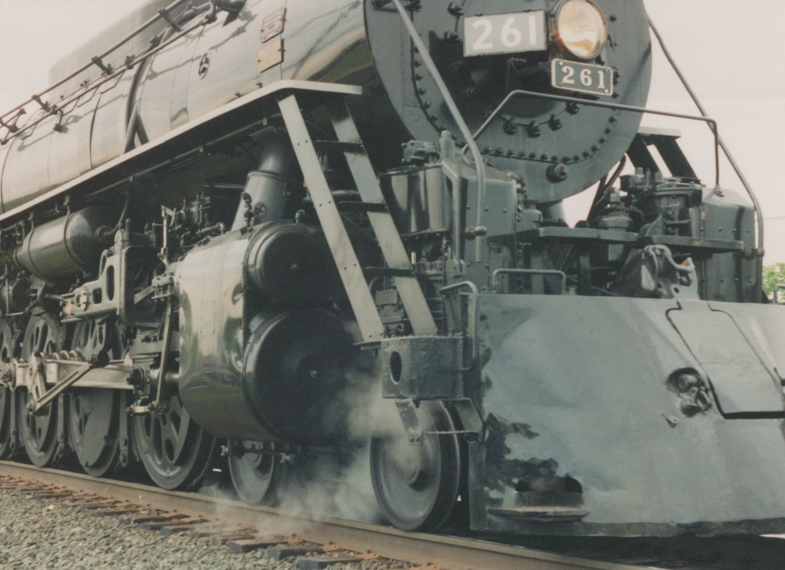A close-up, three-quarter front view of the dark green CB&Q steam locomotive number 261, showing its massive wheels and front details, with steam rising from the track.