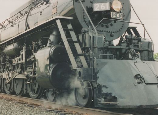 A close-up, three-quarter front view of the dark green CB&Q steam locomotive number 261, showing its massive wheels and front details, with steam rising from the track.