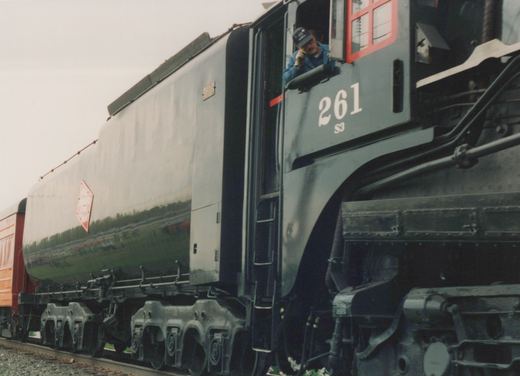Side view of a black steam locomotive, number 261, with a red diamond herald on its side, stopped on tracks with a crew member leaning out of the cab window.