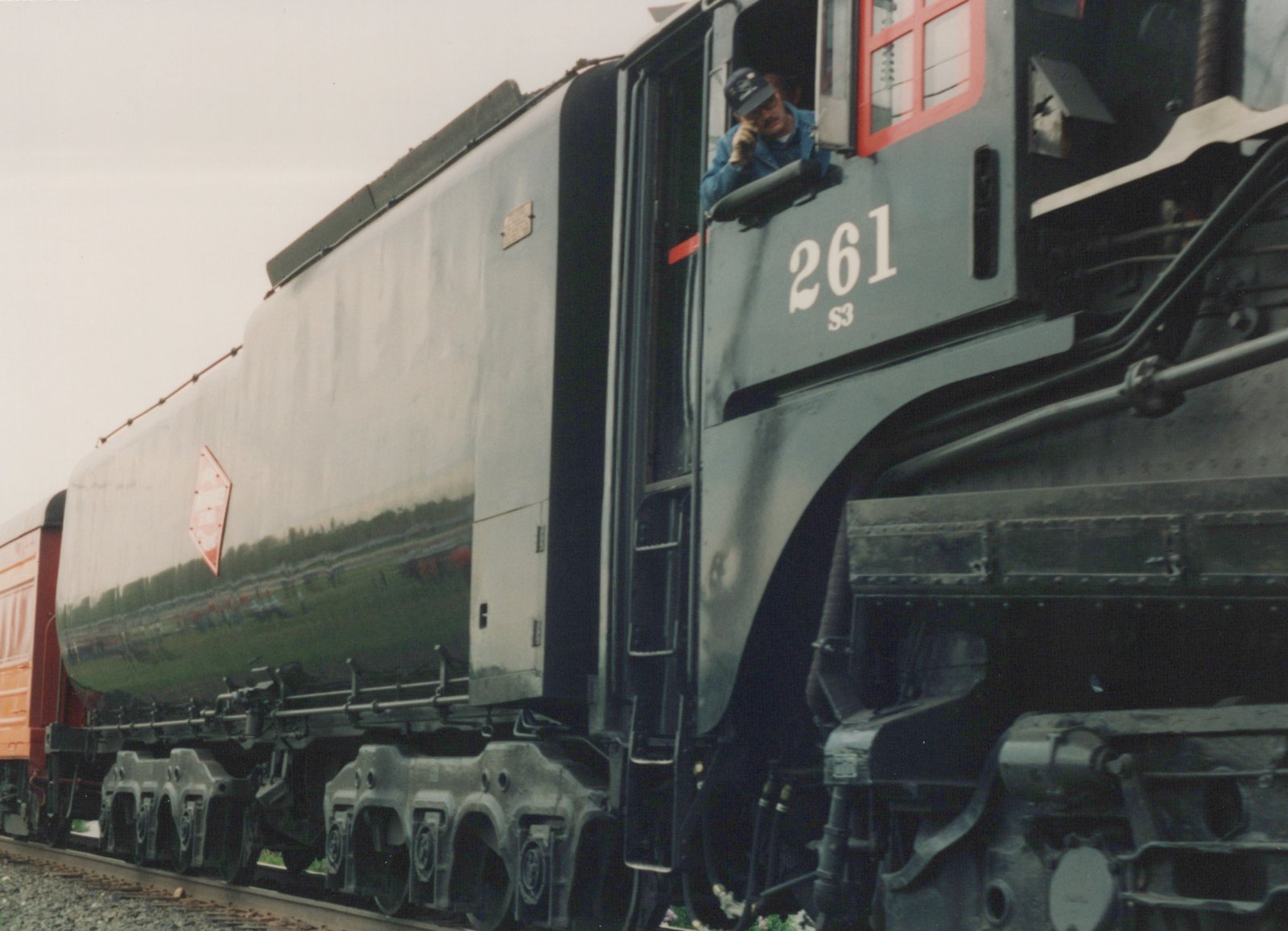 Side view of a black steam locomotive, number 261, with a red diamond herald on its side, stopped on tracks with a crew member leaning out of the cab window.