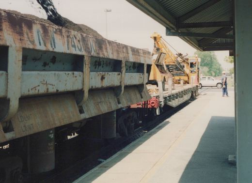 A side view of a rusted Chicago and North Western freight car next to a work train with a yellow crane on a sunny day at a station platform.
