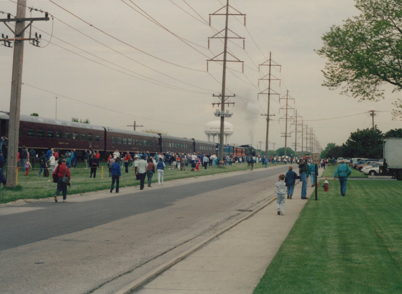A long passenger train led by a black steam locomotive with an orange band travels down a track lined with a large crowd of spectators on a cloudy day, with a 'CITY OF DES PLAINES' water tower in the background.