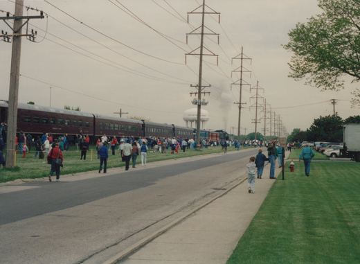 A long passenger train led by a black steam locomotive with an orange band travels down a track lined with a large crowd of spectators on a cloudy day, with a 'CITY OF DES PLAINES' water tower in the background.