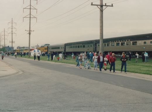 A long, silver passenger train with Bluewater lettering is stopped on a grassy track, surrounded by a large crowd of people walking and standing on the adjacent road and grass under an overcast sky.