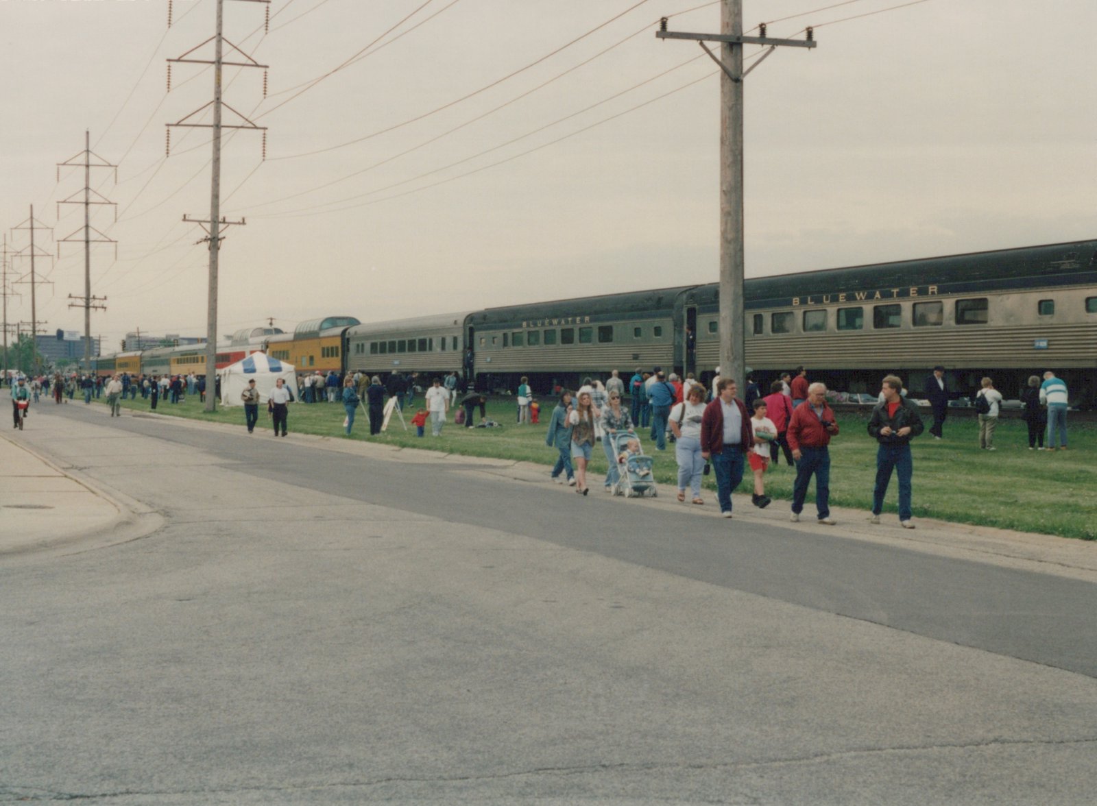 A long, silver passenger train with Bluewater lettering is stopped on a grassy track, surrounded by a large crowd of people walking and standing on the adjacent road and grass under an overcast sky.
