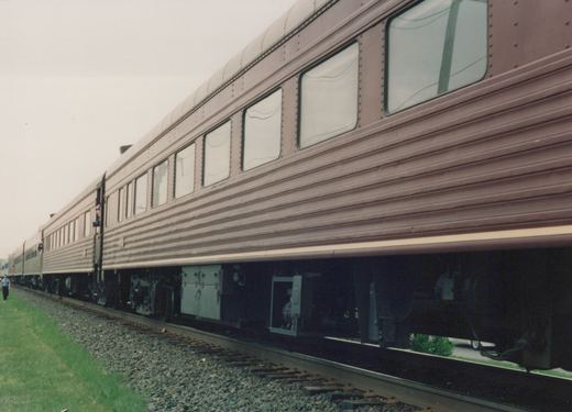 A side view of a maroon and cream Metra gallery car on a gravel-track right-of-way, with a person walking alongside.