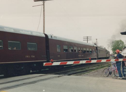 A maroon and silver passenger train with the numbers 716 and 715 crosses a street at a grade crossing while several people, including a child with a bicycle, watch from the side.