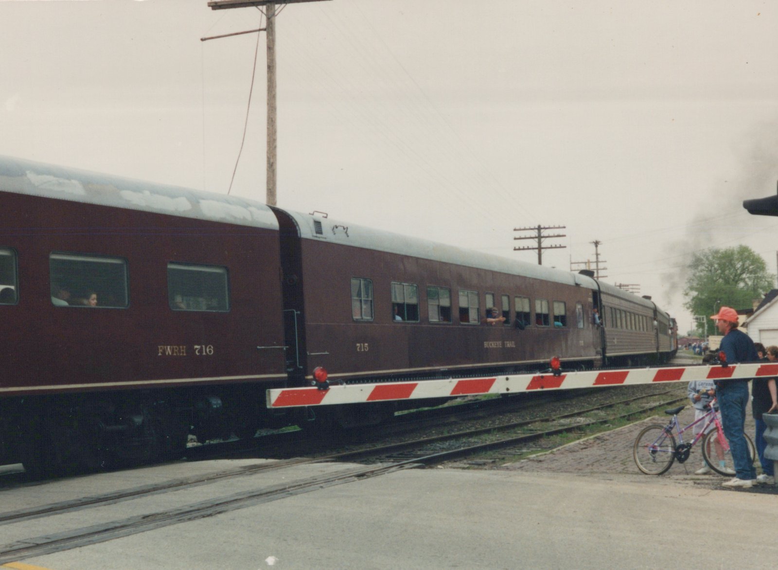 A maroon and silver passenger train with the numbers 716 and 715 crosses a street at a grade crossing while several people, including a child with a bicycle, watch from the side.