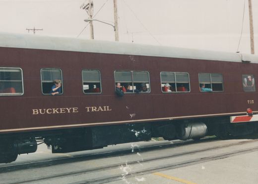 Side view of a maroon passenger railcar with 'BUCKEYE TRAIL' on its side and the number 715 on its end, stopped on tracks in a street with several people visible in the windows.