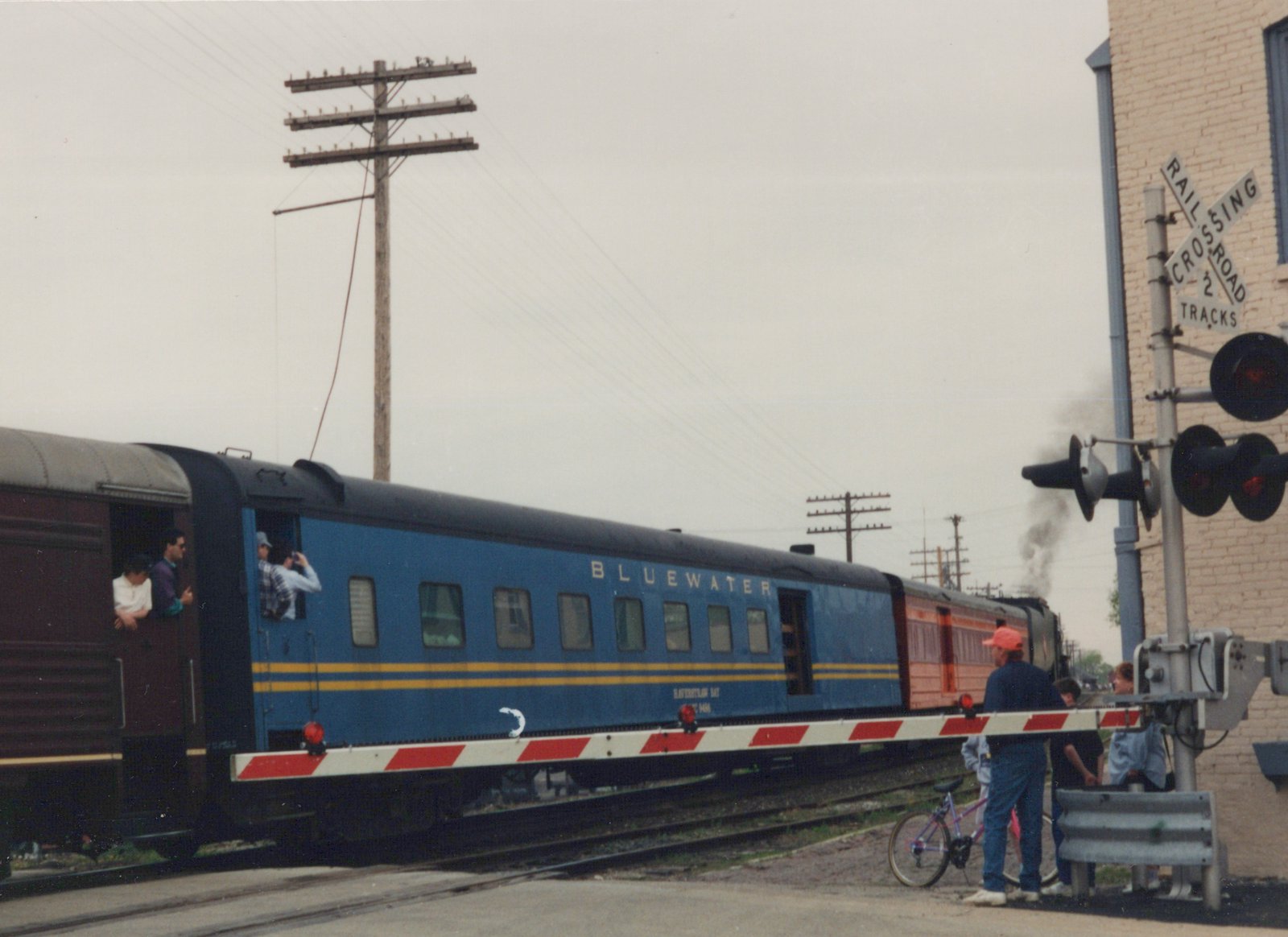 Side view of a long blue and yellow passenger train with 'BLUEWATER' lettering crossing a street; people stand nearby with a bicycle, and a sign reads 'RAILROAD CROSSING 2 TRACKS'.