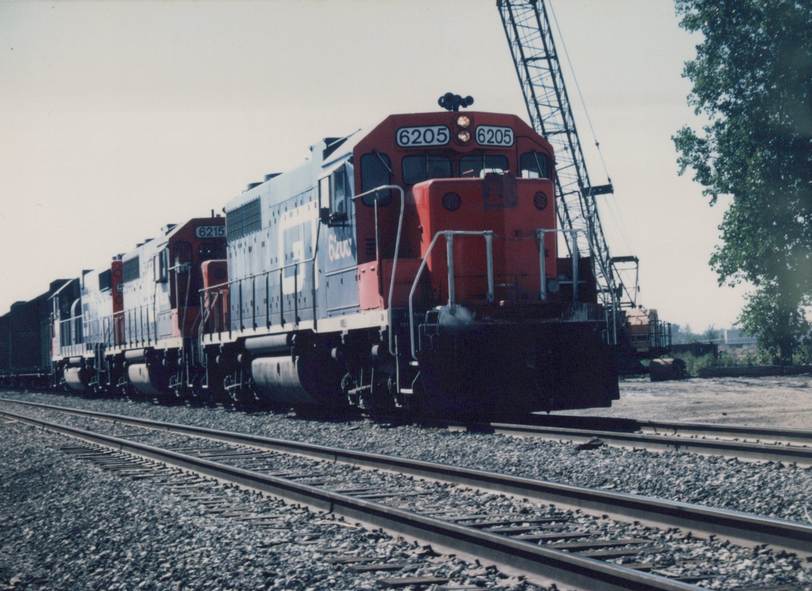 A red and blue Soo Line diesel locomotive, number 6205, is positioned at the front of a train in a railroad yard with a large crane visible in the background.