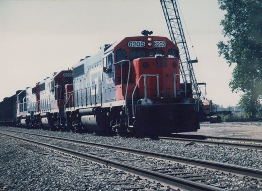 A red and blue Soo Line diesel locomotive, number 6205, is positioned at the front of a train in a railroad yard with a large crane visible in the background.