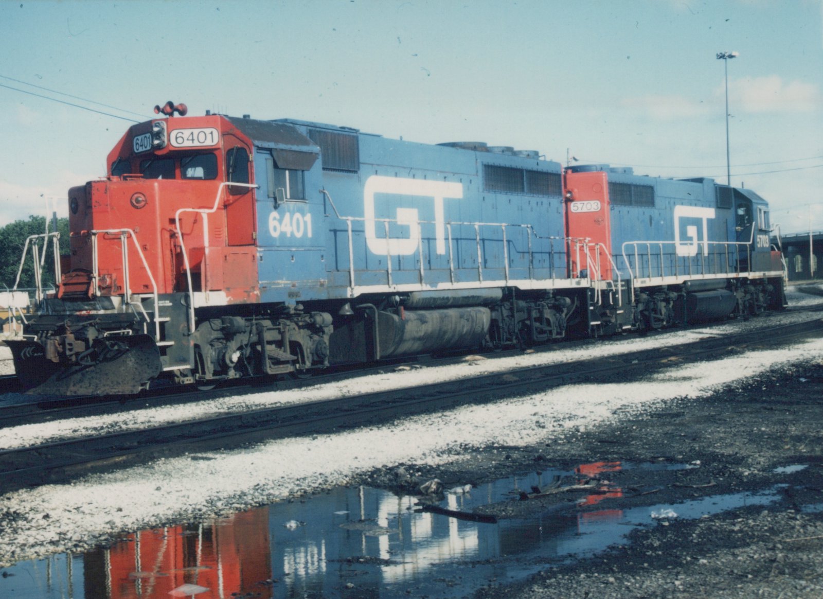 Side view of two blue and red Grand Trunk Western locomotives, numbers 6401 and 5703, parked on tracks with puddles reflecting their colors.