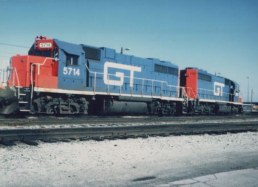 Two blue and red diesel locomotives with 'GT' lettering, numbers 5714 and 5921, are parked on parallel tracks in a snowy yard under a clear blue sky.