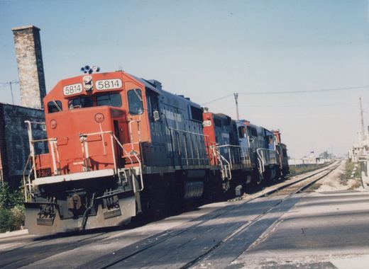 A red and gray Chicago and North Western diesel locomotive, number 5814, leads a train on tracks running through a paved street, with a brick building and smokestack in the background.