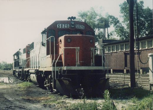A red and gray diesel locomotive with number 5825 on the front, parked in a rail yard with another similar locomotive behind it, surrounded by trees and a wooden building.