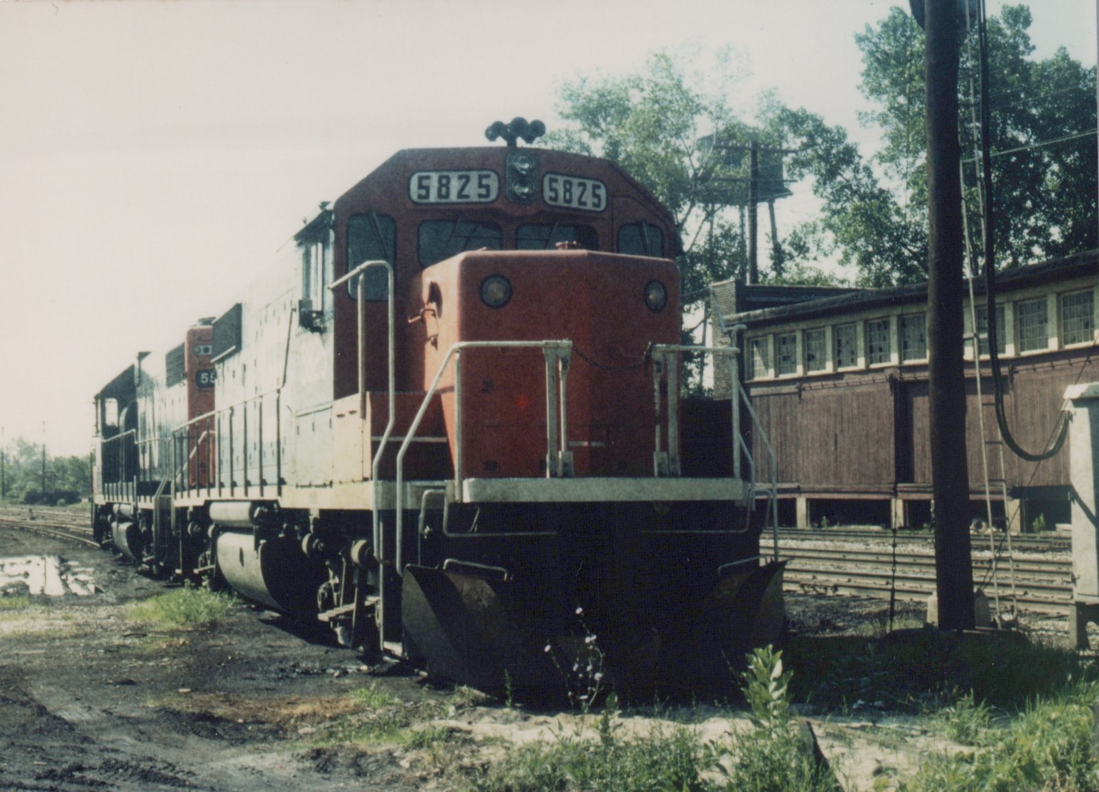 A red and gray diesel locomotive with number 5825 on the front, parked in a rail yard with another similar locomotive behind it, surrounded by trees and a wooden building.
