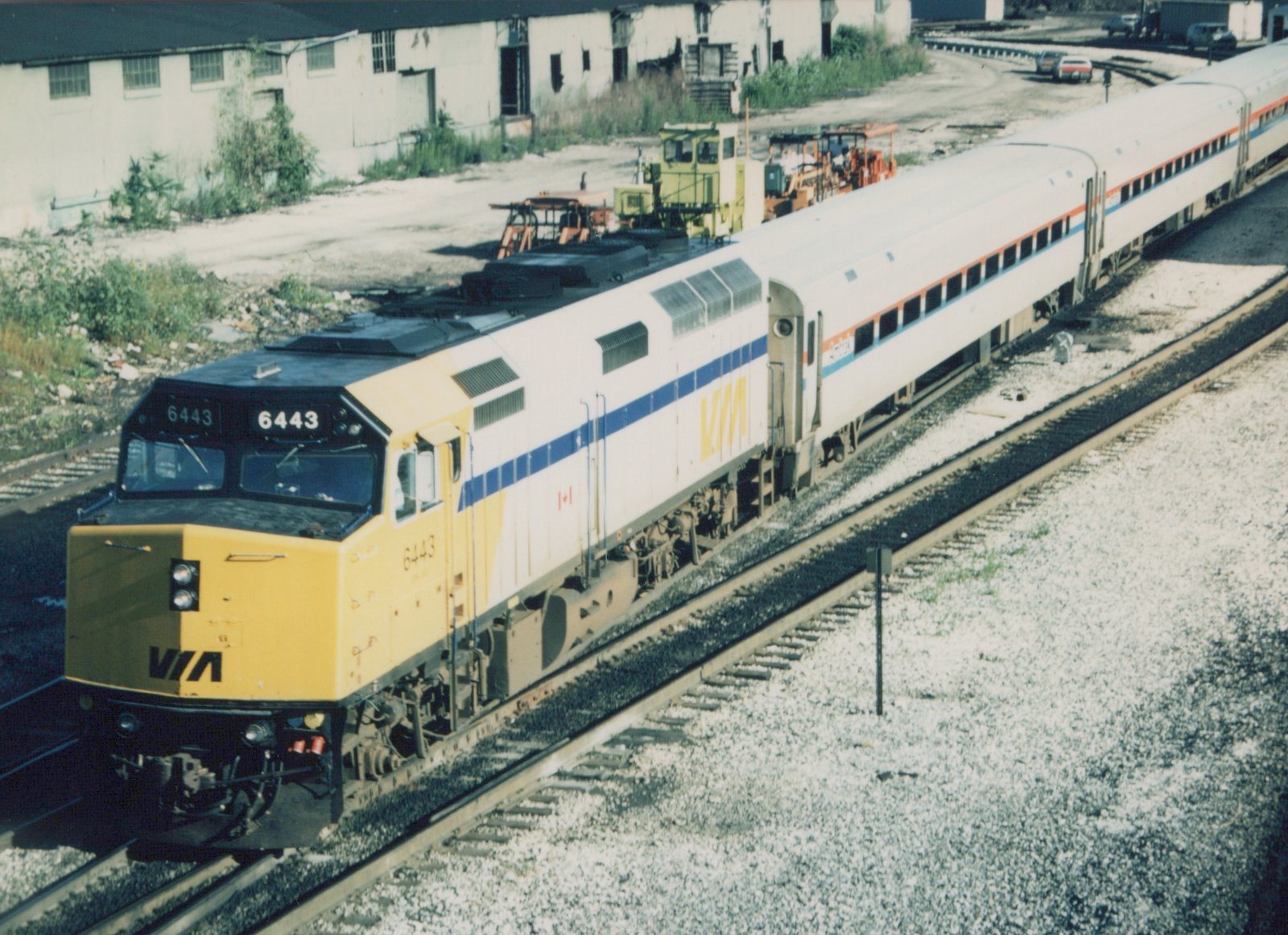 A yellow-nosed VIA Rail Canada diesel locomotive, numbered 6443, pulls a train of white passenger cars with blue and orange stripes along a set of railroad tracks on a sunny day.