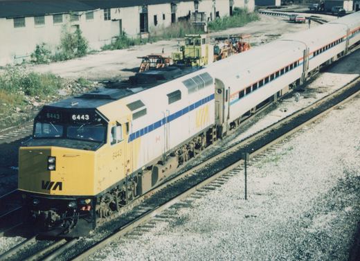 A yellow-nosed VIA Rail Canada diesel locomotive, numbered 6443, pulls a train of white passenger cars with blue and orange stripes along a set of railroad tracks on a sunny day.
