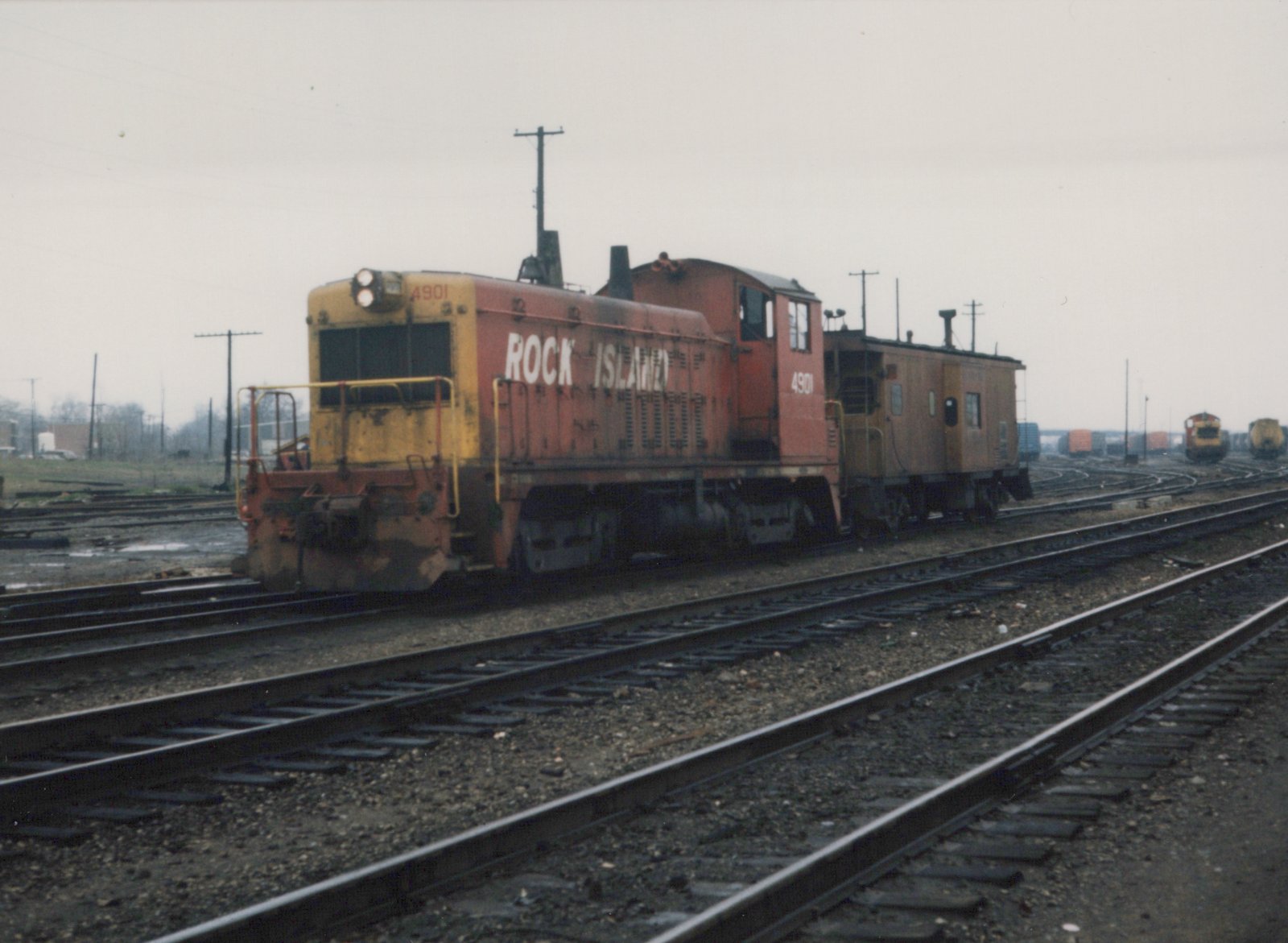 A Rock Island diesel locomotive, number 4901, painted in orange and yellow, is coupled to a matching caboose on a set of tracks in a rail yard with other trains visible in the background.