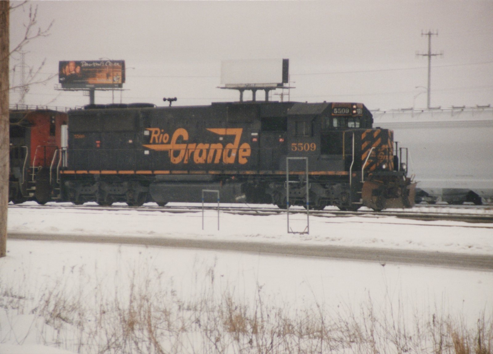 A black and orange diesel locomotive with 'Rio Grande' lettering and the number 5509 is on a snow-covered railroad yard, with a billboard for 'Dawson's Creek' visible in the background.