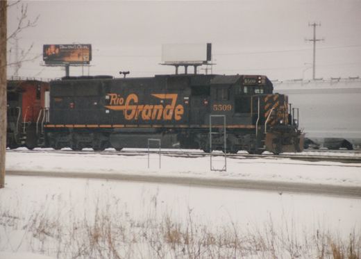 A black and orange diesel locomotive with 'Rio Grande' lettering and the number 5509 is on a snow-covered railroad yard, with a billboard for 'Dawson's Creek' visible in the background.