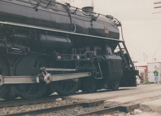 A close-up of a large black steam locomotive with a white stripe and a circular 'M' herald on its tender, parked on tracks in a yard with a person in the background taking a picture.