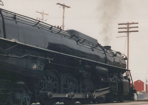 A large black steam locomotive with a 4-8-4 wheel arrangement is parked in a rail yard, emitting smoke from its stack, against a backdrop of utility poles and a red building.