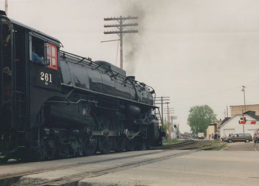 A black steam locomotive with the number 261 on its cab is at a grade crossing, emitting smoke, with spectators and a minivan visible in the background.