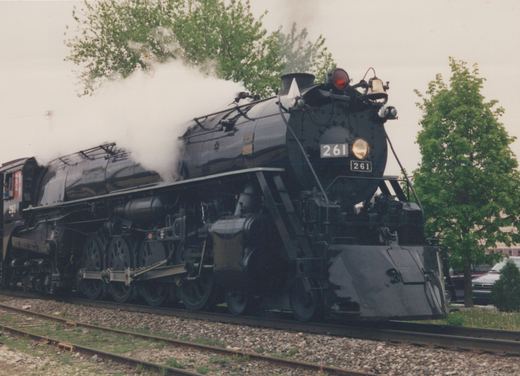 A black steam locomotive numbered 261 moves along a track with steam billowing from its stack, surrounded by green trees and a partially visible automobile in the background.