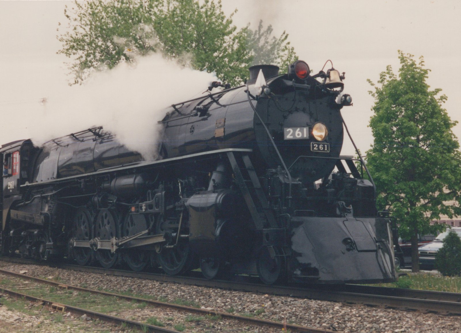A black steam locomotive numbered 261 moves along a track with steam billowing from its stack, surrounded by green trees and a partially visible automobile in the background.