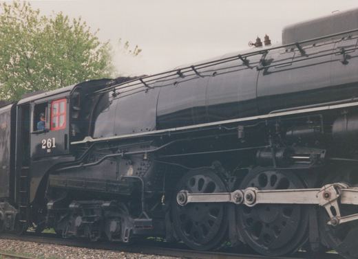 A side view of a large, black steam locomotive with the number 261 on its cab, a person visible in the cab window, and steam venting from the top, set against a backdrop of green trees.