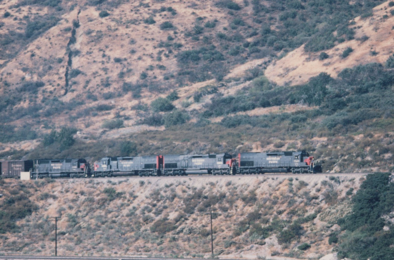 A long freight train with multiple locomotives moves along a track set against a dry, brush-covered hillside under a clear sky.