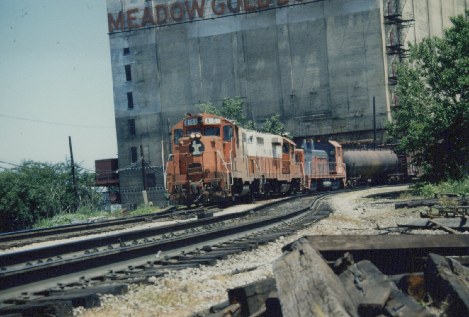 An orange and white Illinois Central diesel locomotive pulls a train on a curved track in front of a large industrial building with 'MEADOW GOLD' lettering.
