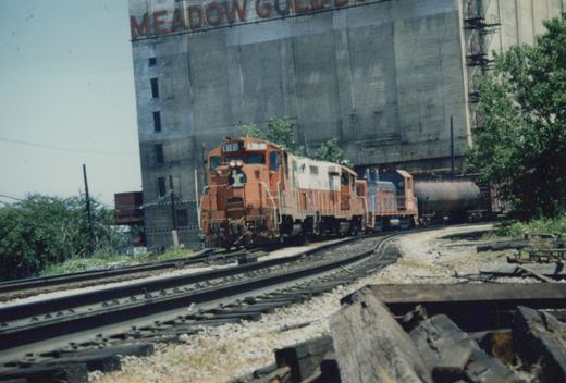 An orange and white Illinois Central diesel locomotive pulls a train on a curved track in front of a large industrial building with 'MEADOW GOLD' lettering.