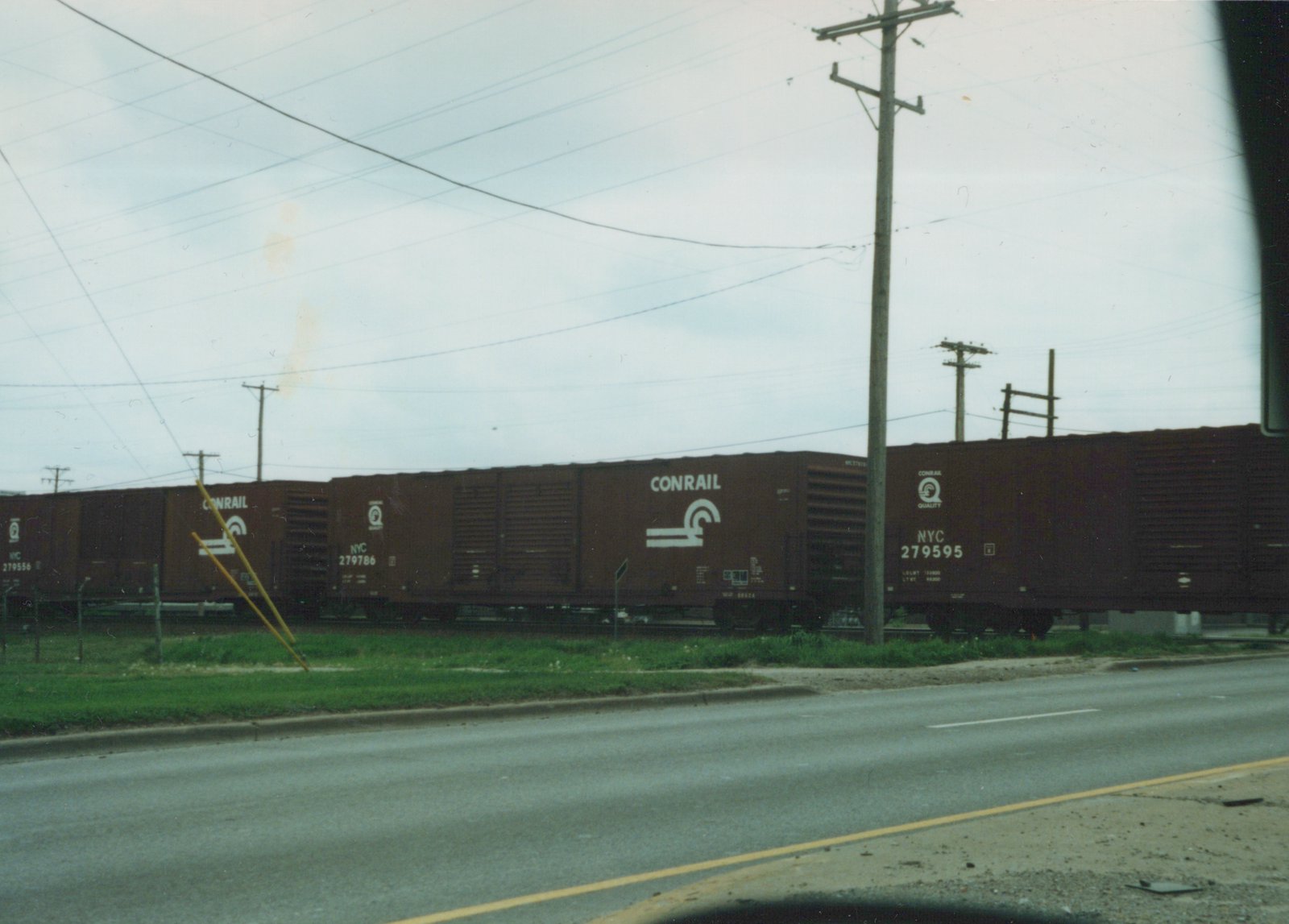 A side view of a brown Conrail freight train with white lettering, including fleet numbers like 279556 and 279595, traveling on tracks alongside a paved road with utility poles and a grassy verge.