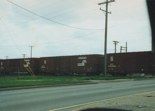 A side view of a brown Conrail freight train with white lettering, including fleet numbers like 279556 and 279595, traveling on tracks alongside a paved road with utility poles and a grassy verge.