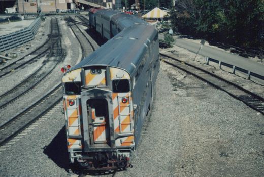 A high-angle view of a Metra commuter train with double-deck gallery cars, showing orange and white chevron markings on the ends, parked in a gravel yard with multiple tracks.