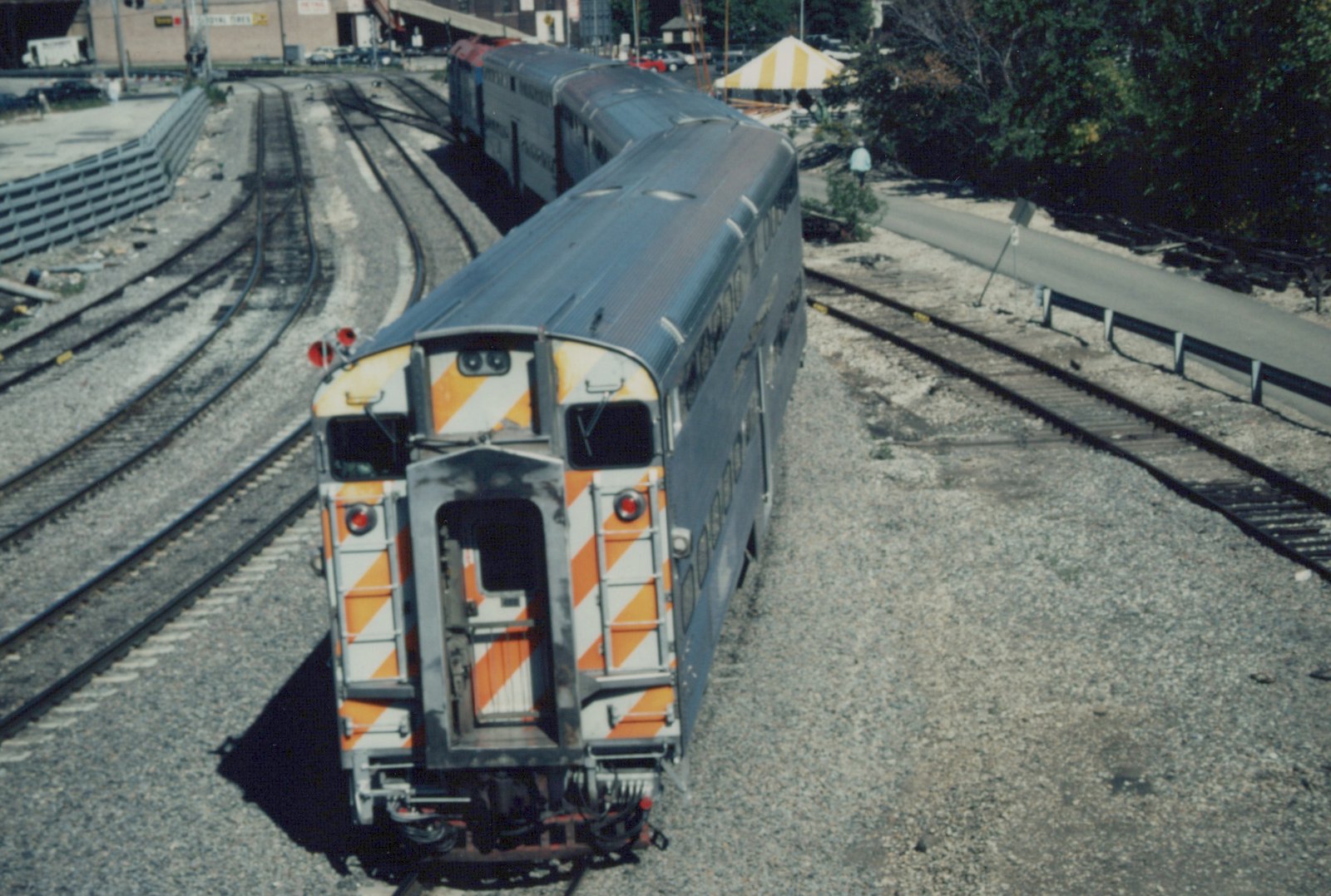 A high-angle view of a Metra commuter train with double-deck gallery cars, showing orange and white chevron markings on the ends, parked in a gravel yard with multiple tracks.